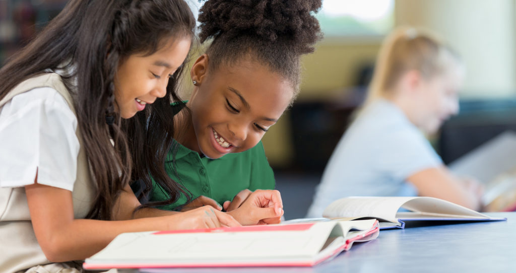Smiling and cheerful schoolgirls reading a book together at school
